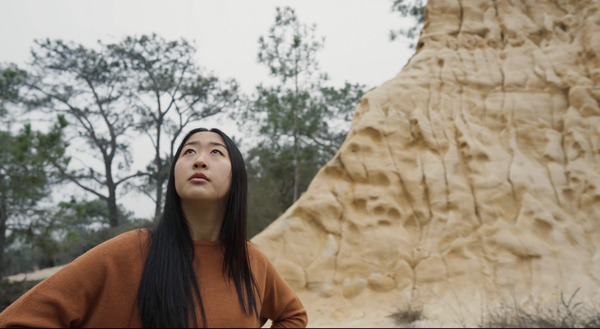 A girl with long black hair and wearing an auburn top looks up at the sky with her hands on her hips. In the background are trees and a tall vertical slab of stone.
