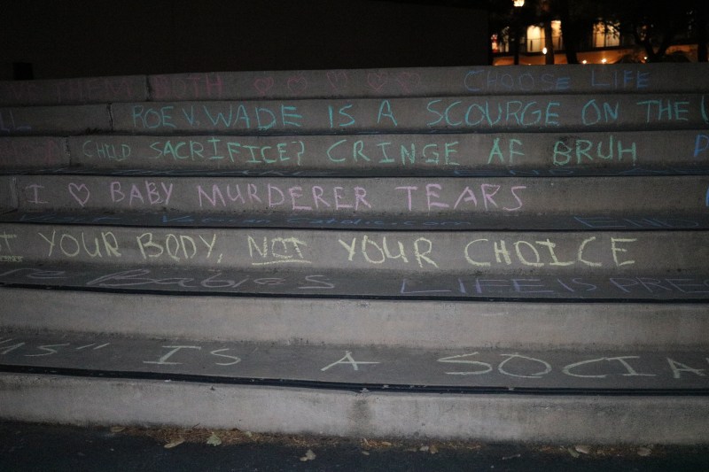 The stairs of White Plaza are decorated with multi-colored chalk. The first visible step reads "Choose life," the second reads "Roe v. Wade is a scourge on the...," the third reads "Child sacrifice? Cringe af bruh," the fourth reads "I love baby murderer tears," and the fifth reads "Your body, not your choice." The charcoal color of the stairs and the dark ambience provides a stark contrast to the colorful chalk.