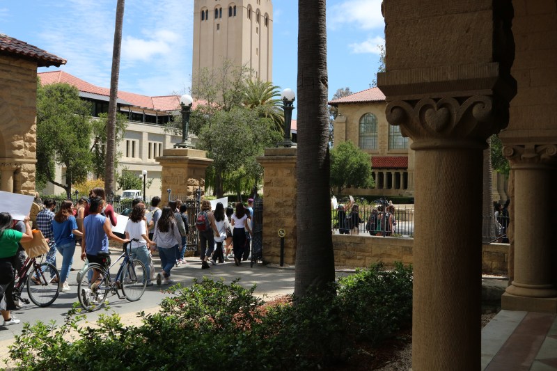 Protestors walk through the gates of Main Quad. Some individuals are walking with their bikes in hand, and many hold posters. Hoover Tower is in the background of the photo.