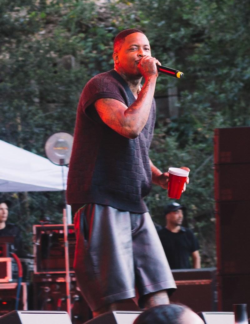Rapper YG performs on stage. He is holding the microphone to his face with his right arm and his holding a solo cup in his left hand.