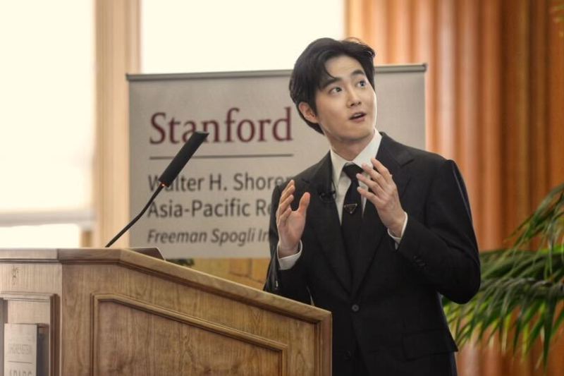 K-Pop artist SUHO stands at a microphone delivering remarks.
