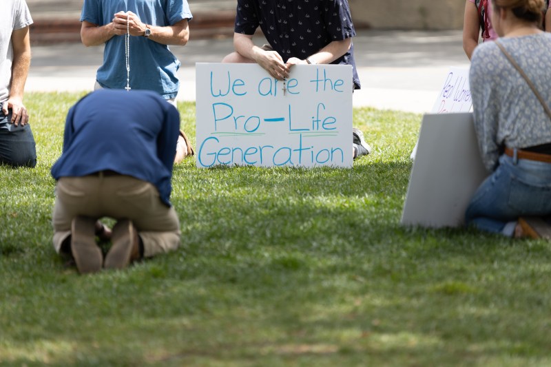 A group of individuals sit in a circle. One person bows their head, while another holds a necklace with a cross. A person sits and holds a sign reading "We are the pro-life generation."