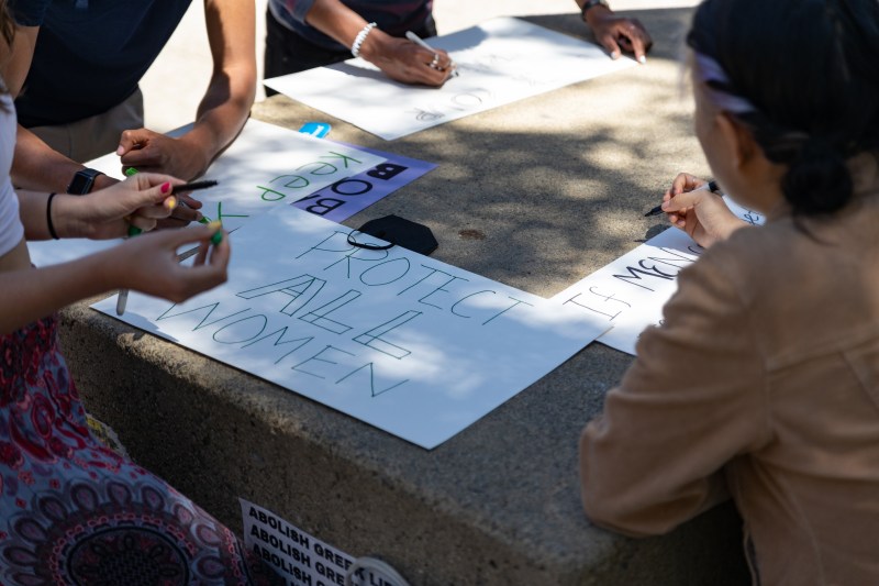 Three attendees surround a concrete table with a poster board and markers in hand. The center poster reads "Protect all women," and the text is written using a green marker.