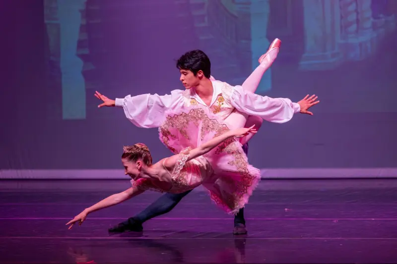 On stage, a male dancer in a loose-fitting shirt strikes a lunge pose while a female dancer hangs horizontally with her legs around his torso, completely off the ground. Both dancers' arms are outstretched.