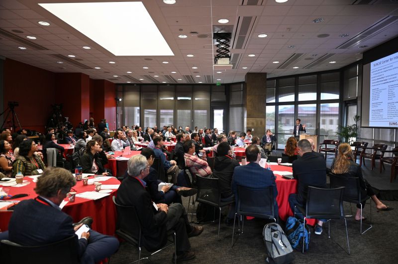 Formally dressed people seated around red tables and facing towards a presentation to the right.