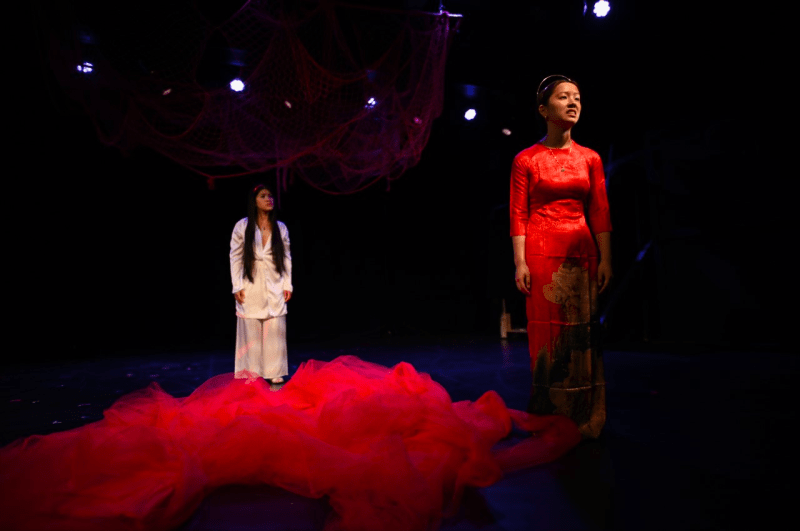 Basked in red light, the person in the front wears a red dress staring longingly into the distance, the person in white dress stands on a pile of red chiffon, glimpses of red nets hangs above them.