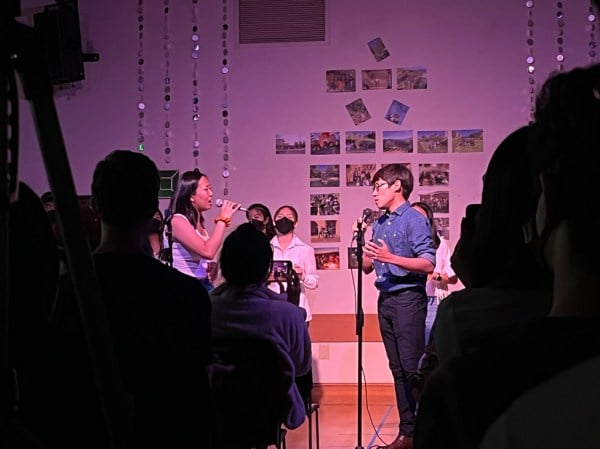 Silhouettes of audience members sit in front of two O-tone singers bathed in a purple and orange light. The two singers solo with microphones in front of the rest of the group.