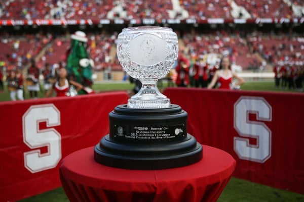 The Director's Cup, a crystal trophy with black base, sits atop a red table with Stanford logos in the background.