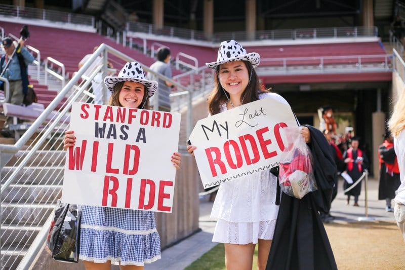 two girls in cow-print cowboy hats hold up signs that read "Stanford was a wild ride" and "my last rodeo"