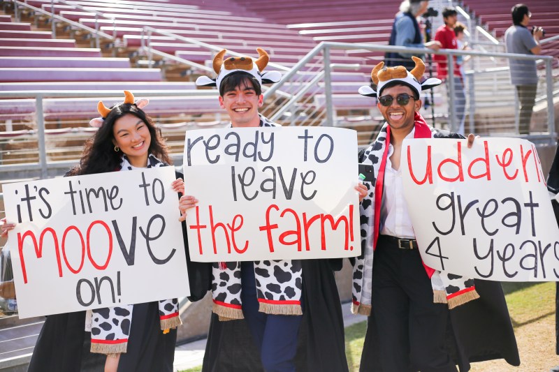Three graduates holding signs that say "it's time to moove on! ready to leave the farm! udderly great 4 years"