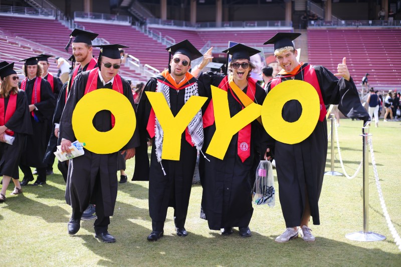 Four graduates in gowns hold up yellow letters "OYYO" to represent the OY/YO art installation.