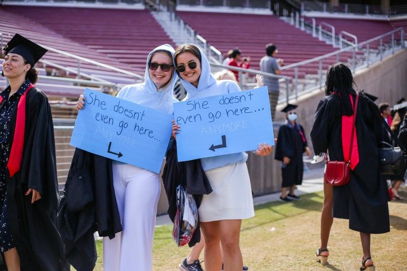 Two graduates hold up signs that say "She doesn't even go here" while dressed in blue hoodies and sunglasses