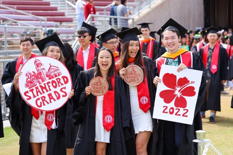 Four Stanford graduates in gowns and caps smile while holding up Trader Joe's cutouts
