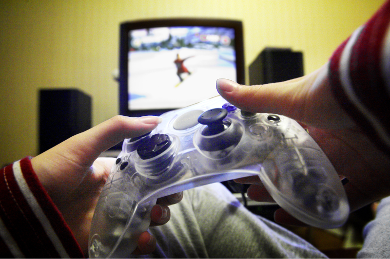 A close up view of a transparent video game controller, held with two hands. In the background is a TV