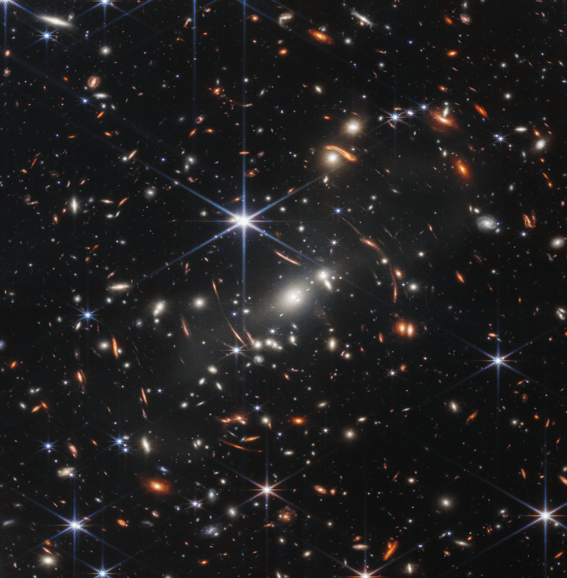 Small orange and white galaxies in oval and spiral shapes are scattered throughout. There are ale blue six-pointed stars in the foreground. Thin orange smudges arc around the center of the image.