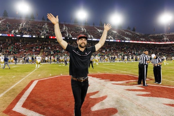 Andrew Luck, dressed in street clothes, throws his hands up while standing in the center of Stanford Stadium.