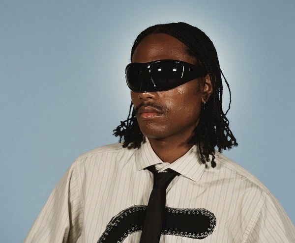 A man [Steve Lacy] looking off to the side with black sunglasses, a white shirt and black tie.