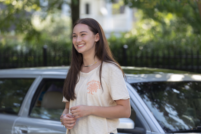 A girl {Belly) standing outside, in front of her car, smiling