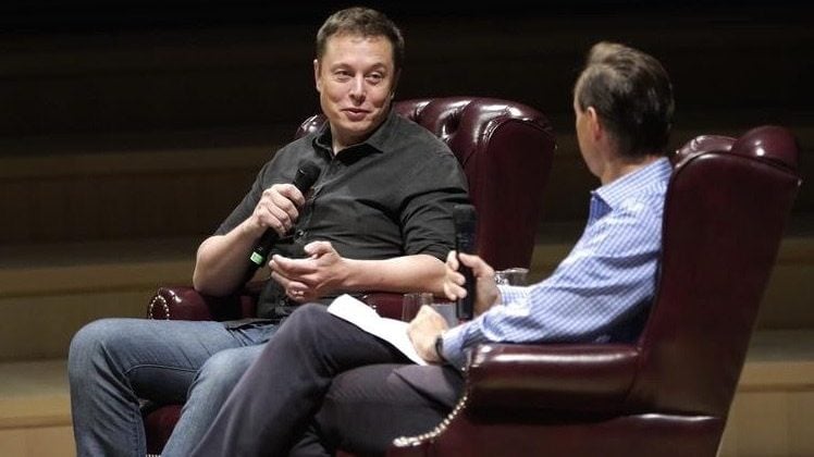 Elon Musk speaks at an interview event, seated in a brown leather armchair with a microphone in hand