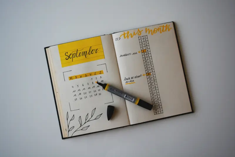An opened journal, with a calendar handwritten on the left page and an uncapped yellow marker on top.