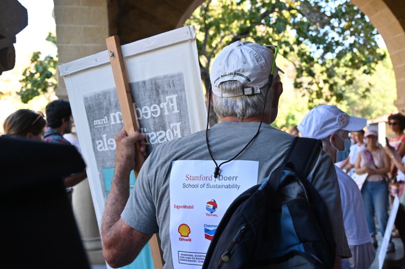 A protestor wears a shirt reading "Stanford School of Sustainability" with logos of gas companies underneath.