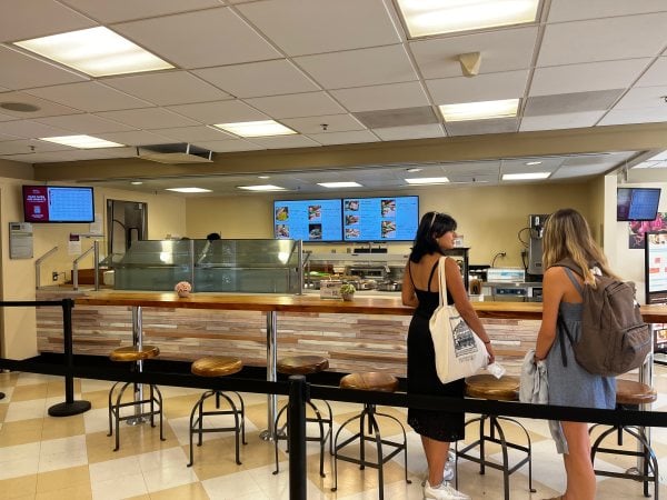 Two people stand in line in front of the Olives counter. A row of barstool chairs in front of them and digital menu displays visible behind the counter.