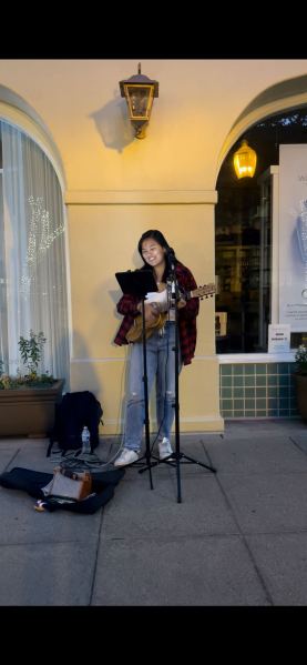 Emma Wong on a sidewalk holding a guitar with a music stand in front of her. An open guitar case to her side.