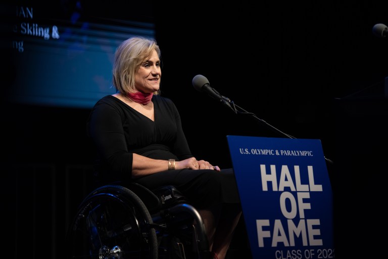 Muffy Davis with a mic and a sign reading "U.S. Olympic and Paralympic Hall of Fame, Class of 2022" in front of her