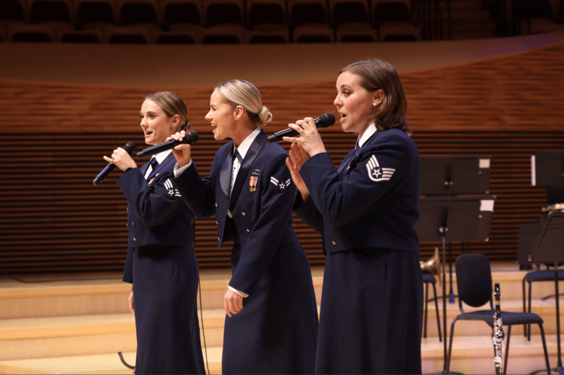 three women in navy blue uniforms stand on a stage and sing into microphones