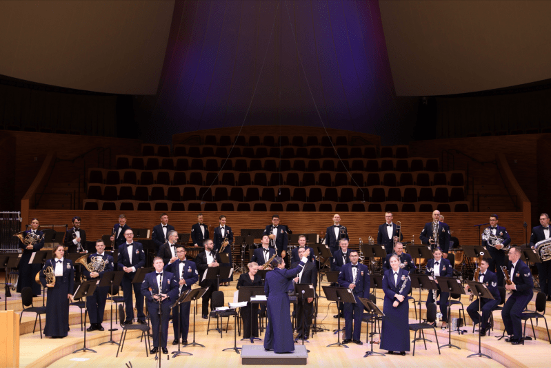 30 band members and a conductor stand on stage, most holding instruments; they are dressed in black and blue uniforms