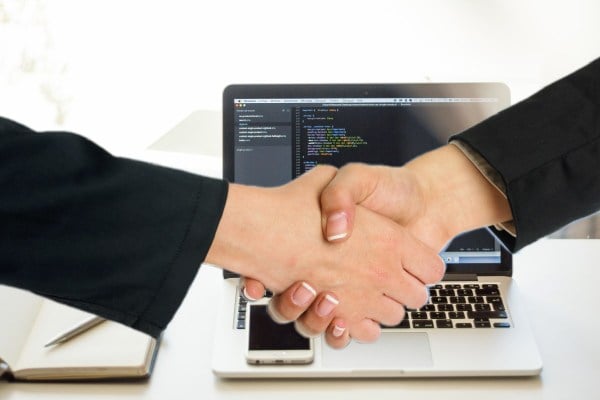 two people in black suits shake hands in front of a laptop