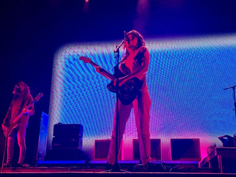 Courtney Barnett playing the guitar and singing into the mic. Blue and red backlight from the big screen fills the stage.