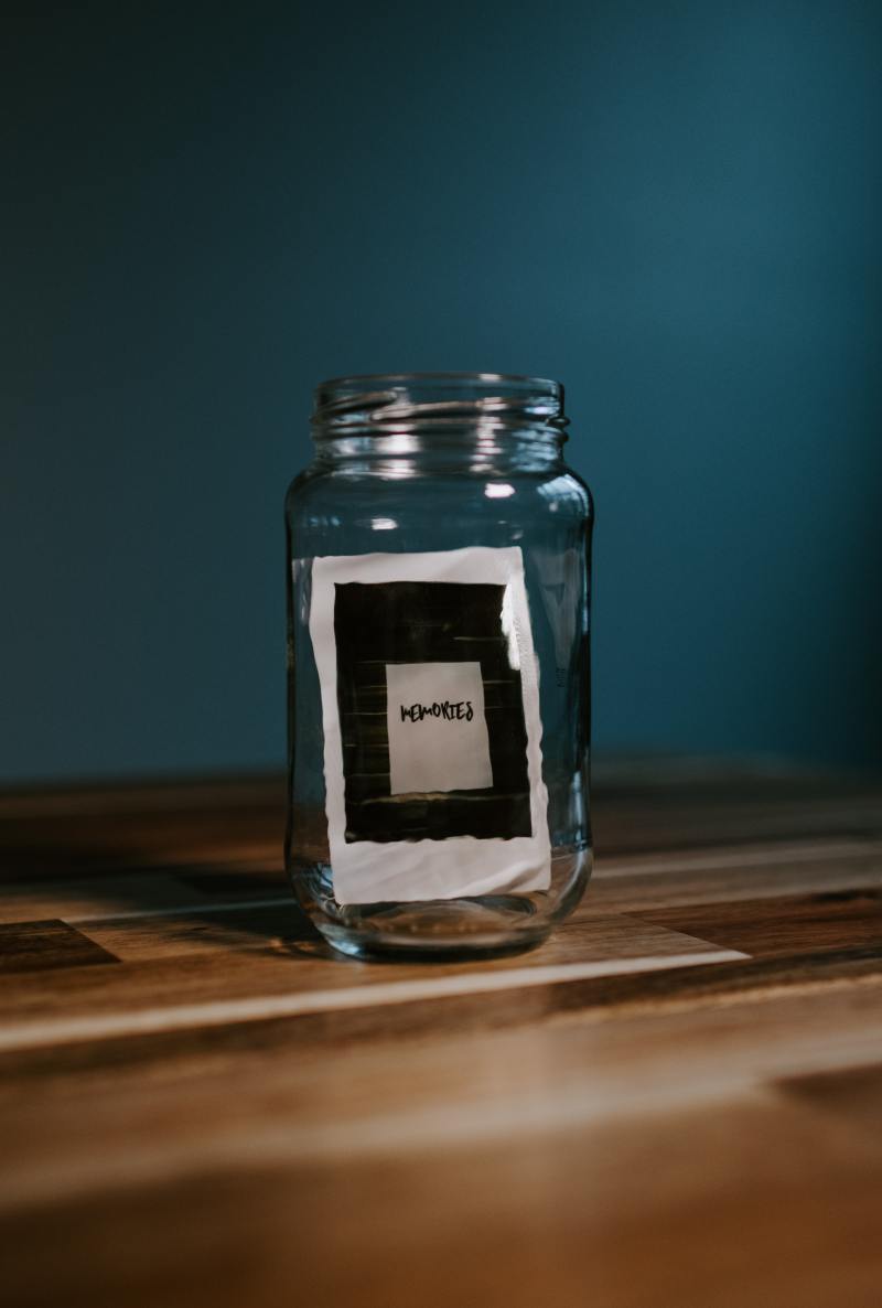 A glass jar with a polaroid photo inside. The polaroid photo is of a white paper against black tiles with "memories" written on it.