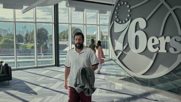 Adam Sandler, bearded, walks in a large glass building with a large Philadelphia 76ers logo statue behind him.