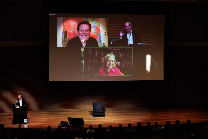 A projector screen shows Zoom boxes featuring Hillary Clinton on the center bottom, Nicholas Kristof in the upper left, and Jim Steyer in the upper right. Some student heads may be in the foreground.