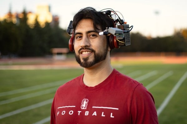 Entrepreneur Kevin Ubilla stands on the track and field arena with his augmented reality headset. In the background is Stanford stadium