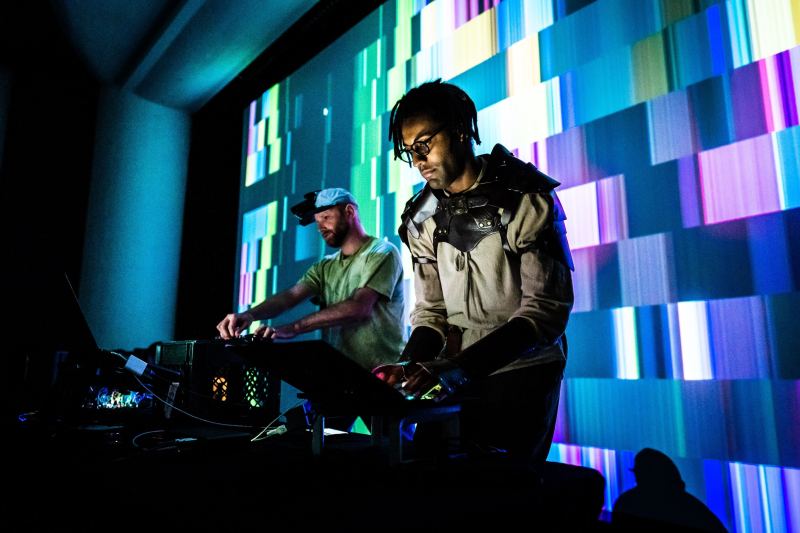 Two people stand in a dark room at a table with sound mixing gear and in front of a glitchy wall light display. The person on the left wears a tee shirt and cap; the one on the right wears a sweatshirt under a black shoulder harness