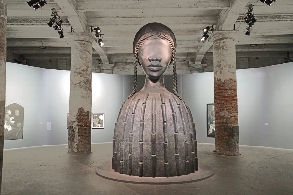 A steel installation of the upperbody of what appears to be a woman without eyes. The background environment contains slightly rusty pillars and grey walls with artworks hung on them.