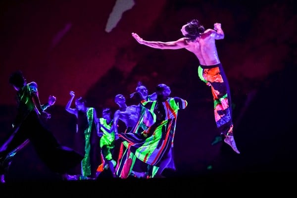 Seven dancers, dressed in colorful neon, dance on the stage. One dancer throws himself up into the air as the others watch intently