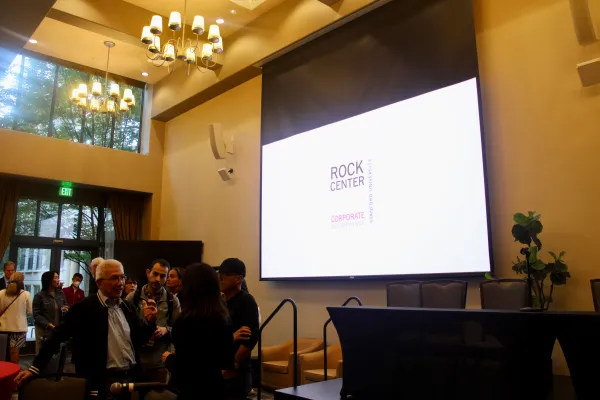 A projector screen reads "Rock Center Corporate Governance Stanford University" above a small stage. On the left is a group of people mingling.
