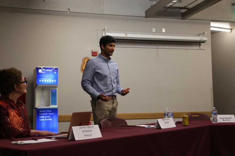 Sathvik Nori stands behind a table with a placard reading his name, a person sitting to his right looks up at him