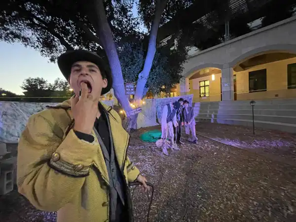 An actor in a yellow coat and hat stares at the screen, pretending to spray something into his mouth. In the background, three haggard-looking actors huddle beneath a tree. A sign on the tree reads: "PRETEND THERE ARE NO LEAVES." Another sign below it reads: "A COUNTRY ROAD, A TREE, EVENING."