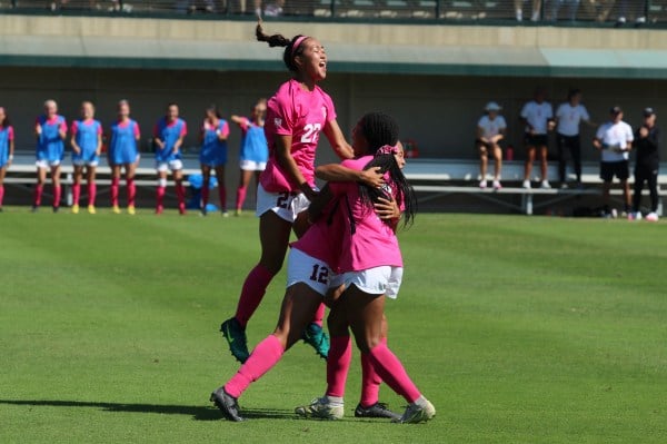 Three Stanford players in pink jerseys celebrate and hug on the field.