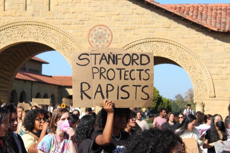 A protester holds up a sign made of cardboard that reads "Stanford Protects Rapists" in Main Quad.
