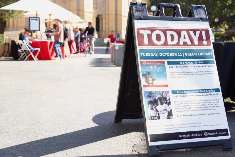 Sign saying "TODAY!" listing events at Green Library on Tuesday, October 11 with red tables and umbrellas in the background.