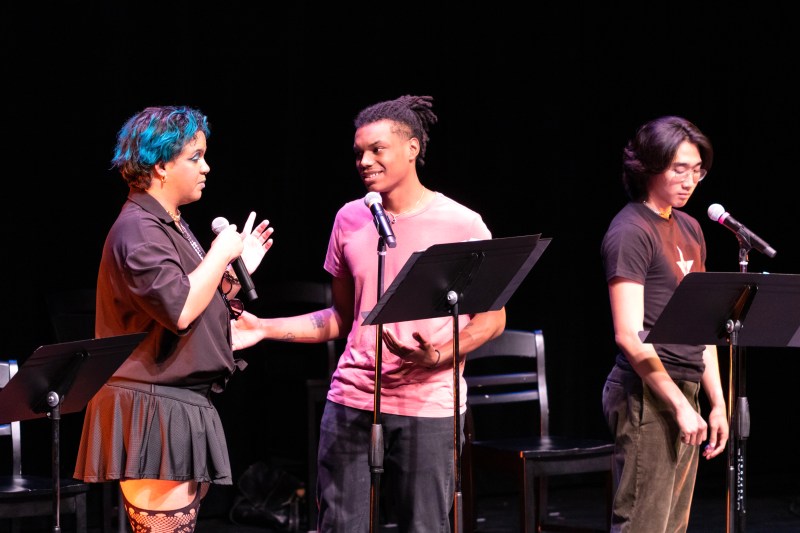 Three people stand on a stage in front of music stands, speaking into microphones. The two leftmost people are gesturing toward each other, while the third speaks toward the audience