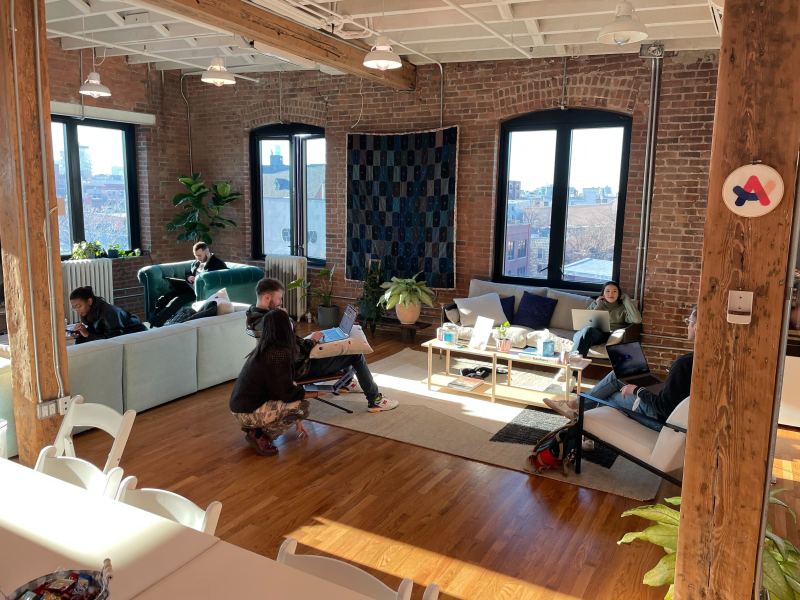 Multiple people with laptops sit working on light-colored furniture in a wooden and brick room illuminated by natural window light and a few overhead light fixtures.
