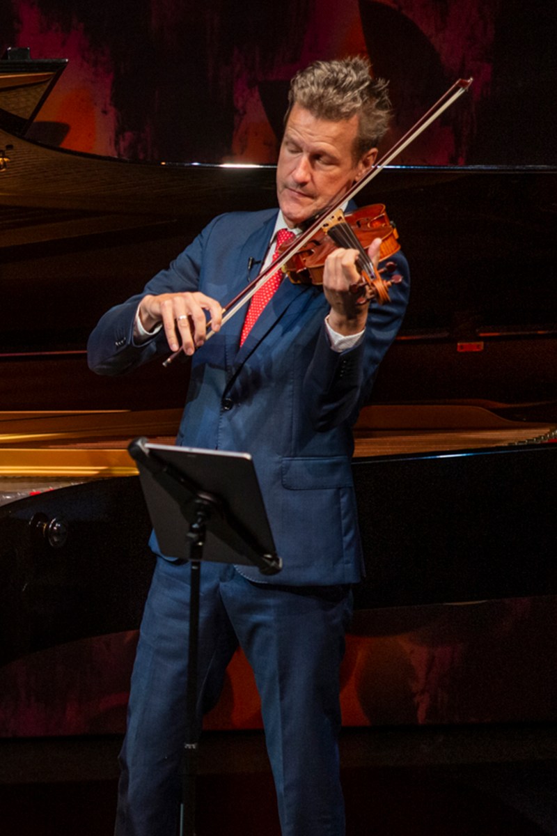 Nuttall plays violin in a blue suit with a white shirt and red tie. His eyes are closed, as if concentrating on the music. A music stand is in front of him.
