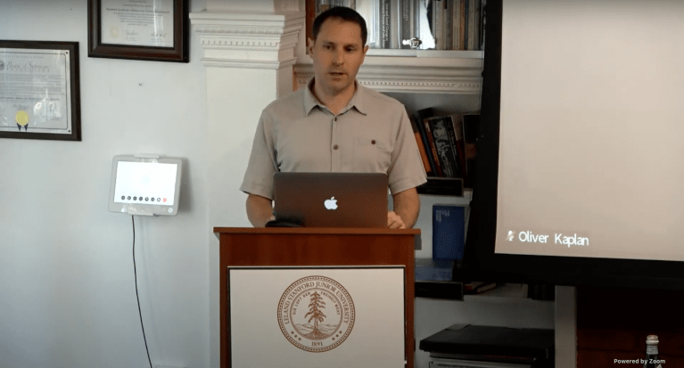 Oliver Kaplan stands at a podium with laptop and speaks to an audience