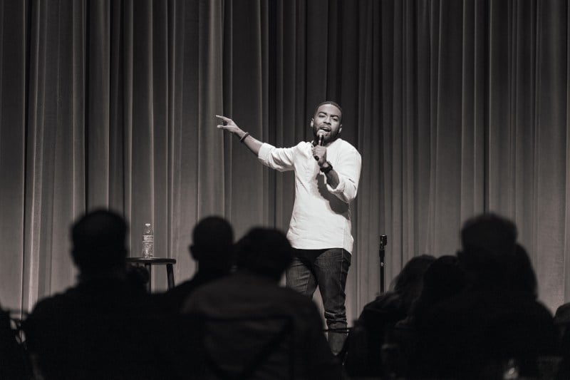 In a black-and-white photo, Ian Lara stands on the stage of Bing Studio holding a mic in his left hand, delivering a monologue to the audience sitting in front of him.